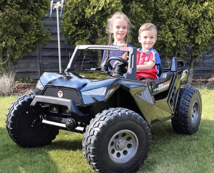 Two kids sit in a Black 24V/4X4 Dune Buggy Ride On UTV on a lawn. The smiling long-haired girl holds the steering wheel, while the boy beside her also smiles. Trees and a wooden fence are visible in the background.