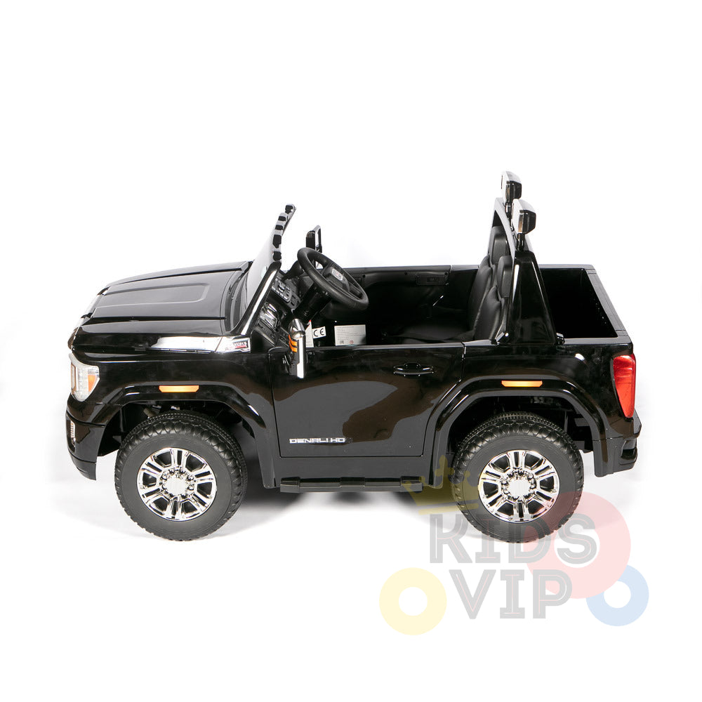 A black 4WD GMC Sierra kids ride-on car with shiny silver wheels, open doors, and a visible steering wheel is shown from the side on a white background. Denali appears on the door. This 2x12V licensed toy provides excitement and parental remote control for added safety.