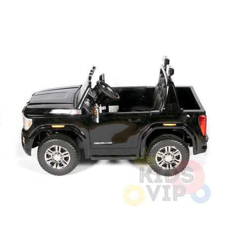 A black 4WD GMC Sierra kids ride-on car with shiny silver wheels, open doors, and a visible steering wheel is shown from the side on a white background. Denali appears on the door. This 2x12V licensed toy provides excitement and parental remote control for added safety.