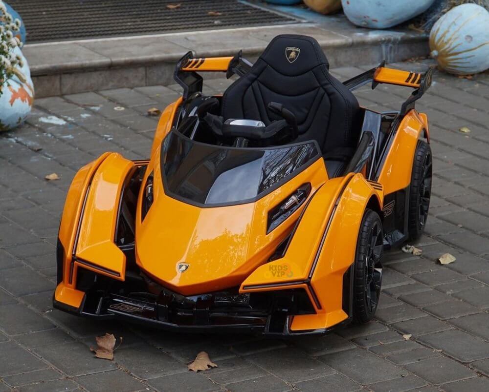 An Orange 12V Official Lamborghini Vision GT kids ride-on car sits on a paved area, featuring a sleek design with black accents and detailed bodywork. Fallen leaves are scattered around it.