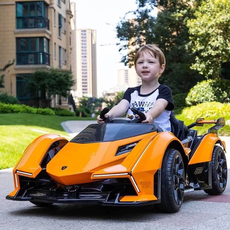 A young child beams while cruising in an Orange 12V Official Lamborghini Vision GT Kids Ride-On Car along a residential path. Tall buildings and greenery create the backdrop as this sleek, battery-powered dream car glides by.
