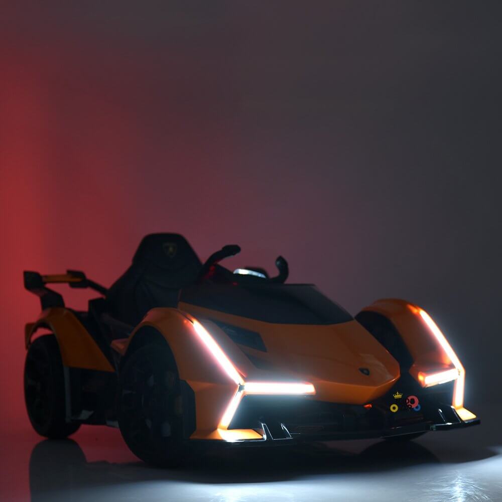 An Orange 12V Official Lamborghini Vision GT Kids Ride-on Car with a bold, angular design and glowing headlights sits on a reflective surface. It features black accents, a wing-like spoiler, and resembles a sleek futuristic toy car against a dark background.