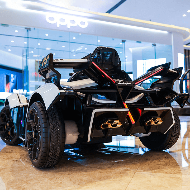 A White 12V Official Lamborghini Vision GT Kids Ride-on Car with a sleek black/white design and prominent rear lights sits indoors on a shiny floor before a store with a bright oppo sign.