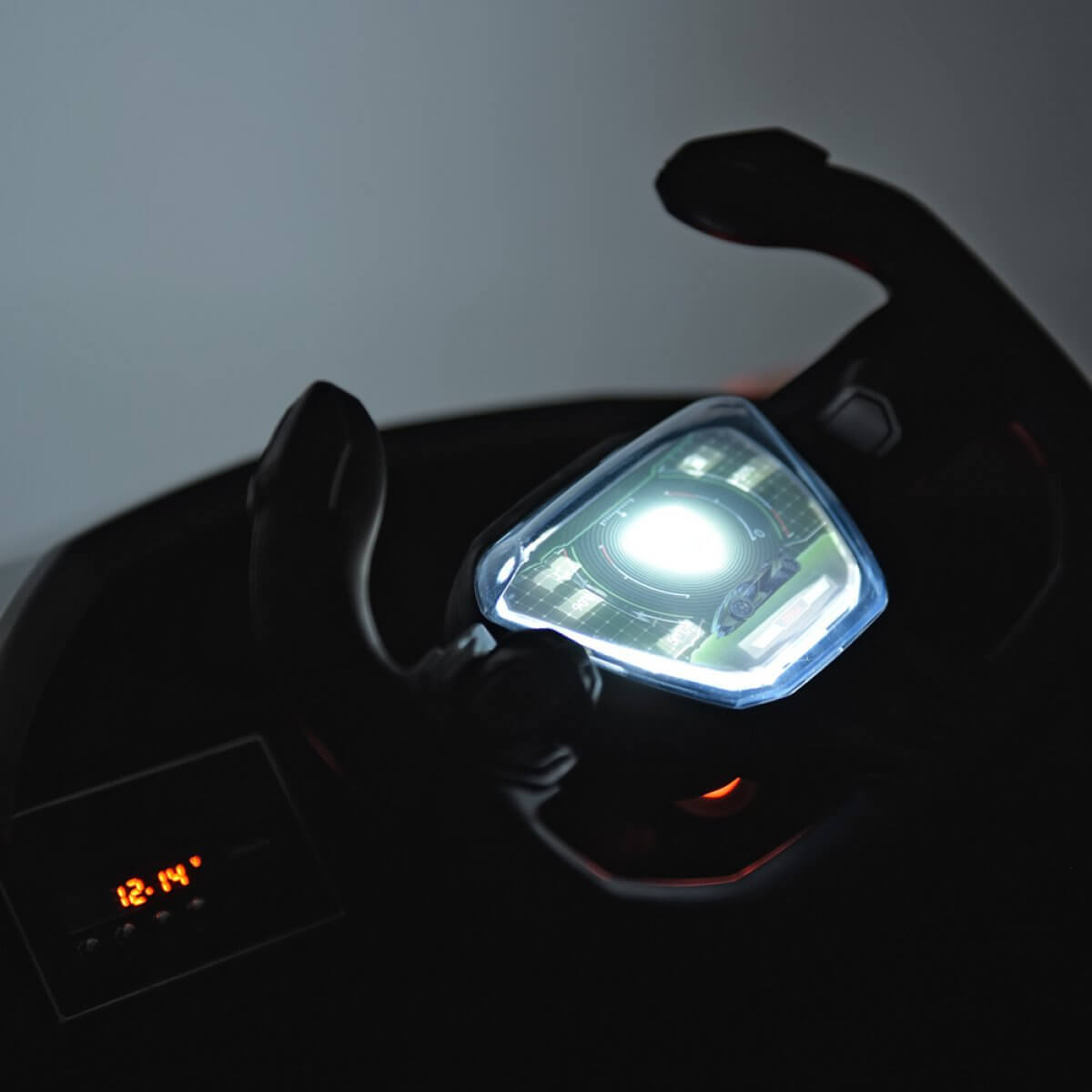 Close-up of an illuminated digital display and steering wheel in the dimly lit Orange 12V Official Lamborghini Vision GT Kids Ride-On Car. The display reads 12:14 in orange, while a bright light reminiscent of the Lamborghini Vision GT shines from the central panel.