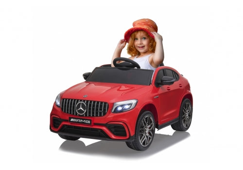 A child with red hair, sporting a red hat, joyfully sits in an Enhance Your Driving Experience with a 12V Mercedes GLC Remote Control ride-on car. This sleek toy features realistic AMG branding, headlights, and grille details for thrilling adventures.