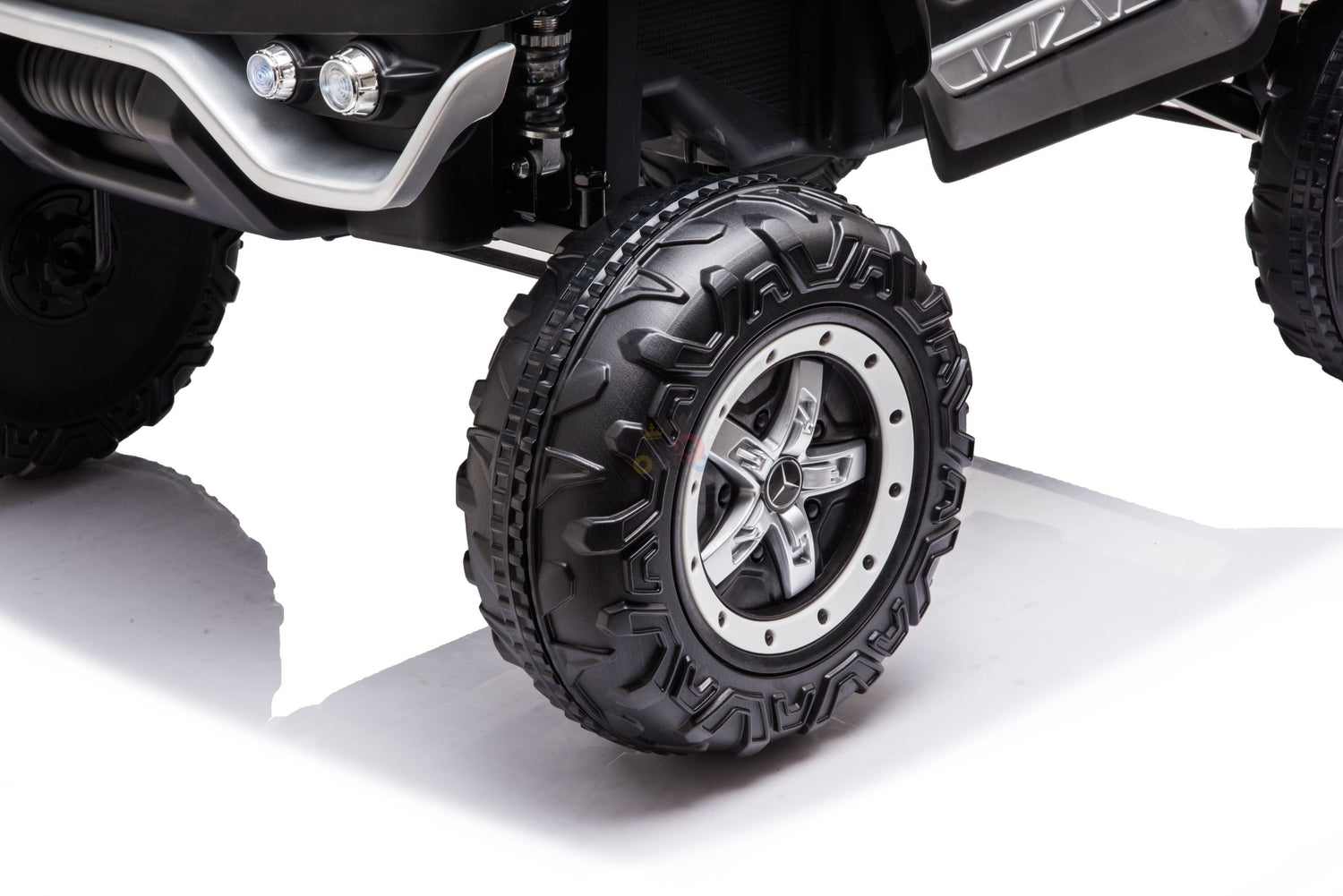 A close-up of the White 12V/4X4 Official Mercedes Unimog Junior Ride On UTV reveals a rugged tire with deep treads, reminiscent of an off-road vehicle. Its metallic rim features a five-spoke design, and the kids ride-on boasts visible suspension and a sleek black exterior with a polished chrome bumper.