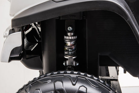 Close-up of the suspension and tire on a White 12V/4X4 Official Mercedes Unimog Junior Ride On UTV. The coiled spring, shock absorber, and rugged tire tread emphasize durability and performance.