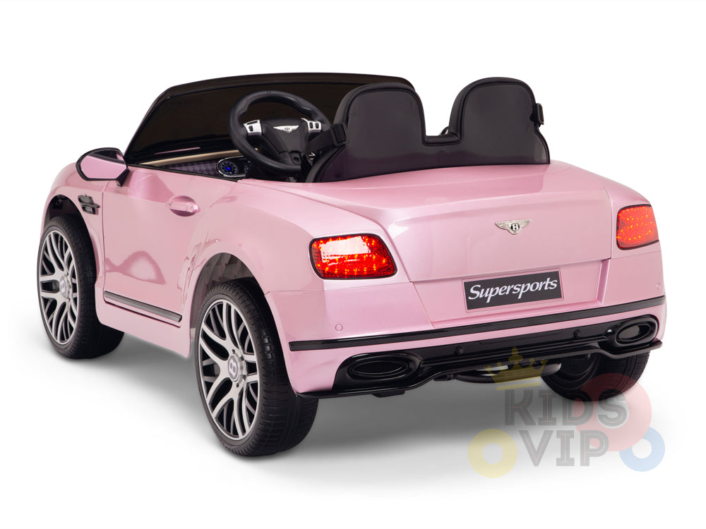 The Pink 12V Bentley GTC Kids Licensed Ride On Car, featuring rubber wheels and parental remote control for safety, resembles a Bentley Continental Sport. It has two seats, a steering wheel, realistic details like rear lights and rims, is shown from behind with a Supersports logo.