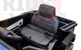 Inside the cabin of the Futuristic 12V Police Truck Ride On for Kids and Toddlers, a black leather seat with stitching detail and a seatbelt is visible. The steering wheel appears partially on the left, set against faint colorful text in the background.