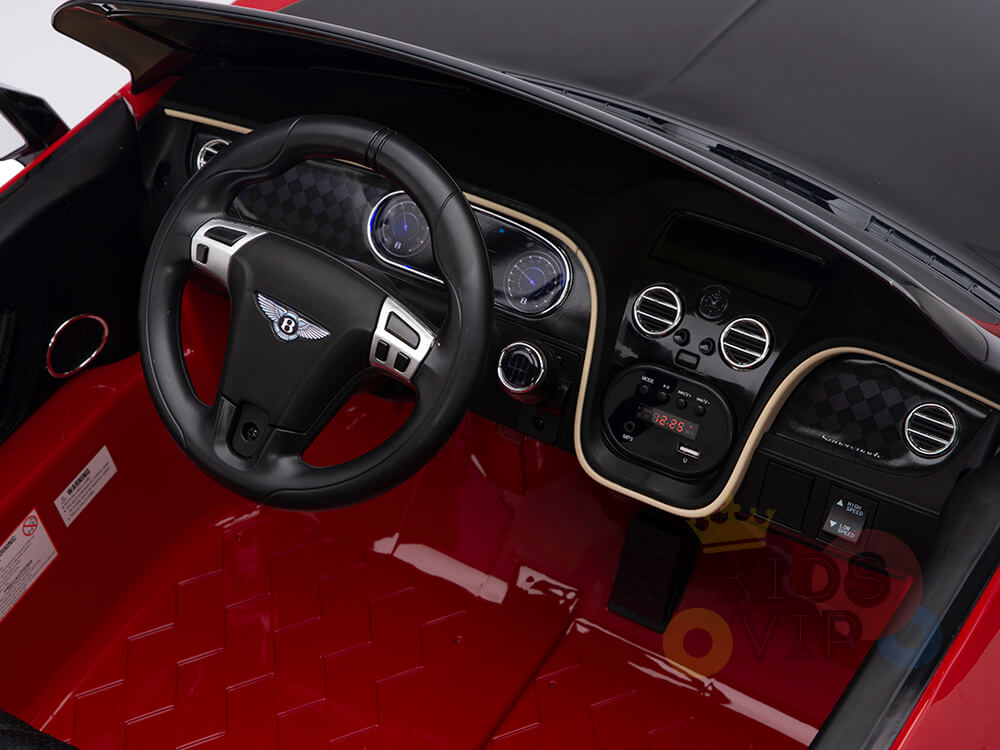The Red 12V Bentley GTC Kids Licensed Ride On Car boasts a black steering wheel with metallic accents, a detailed dashboard, and a red footwell. It mimics luxury interiors with realistic knobs and dials for an authentic driving experience.