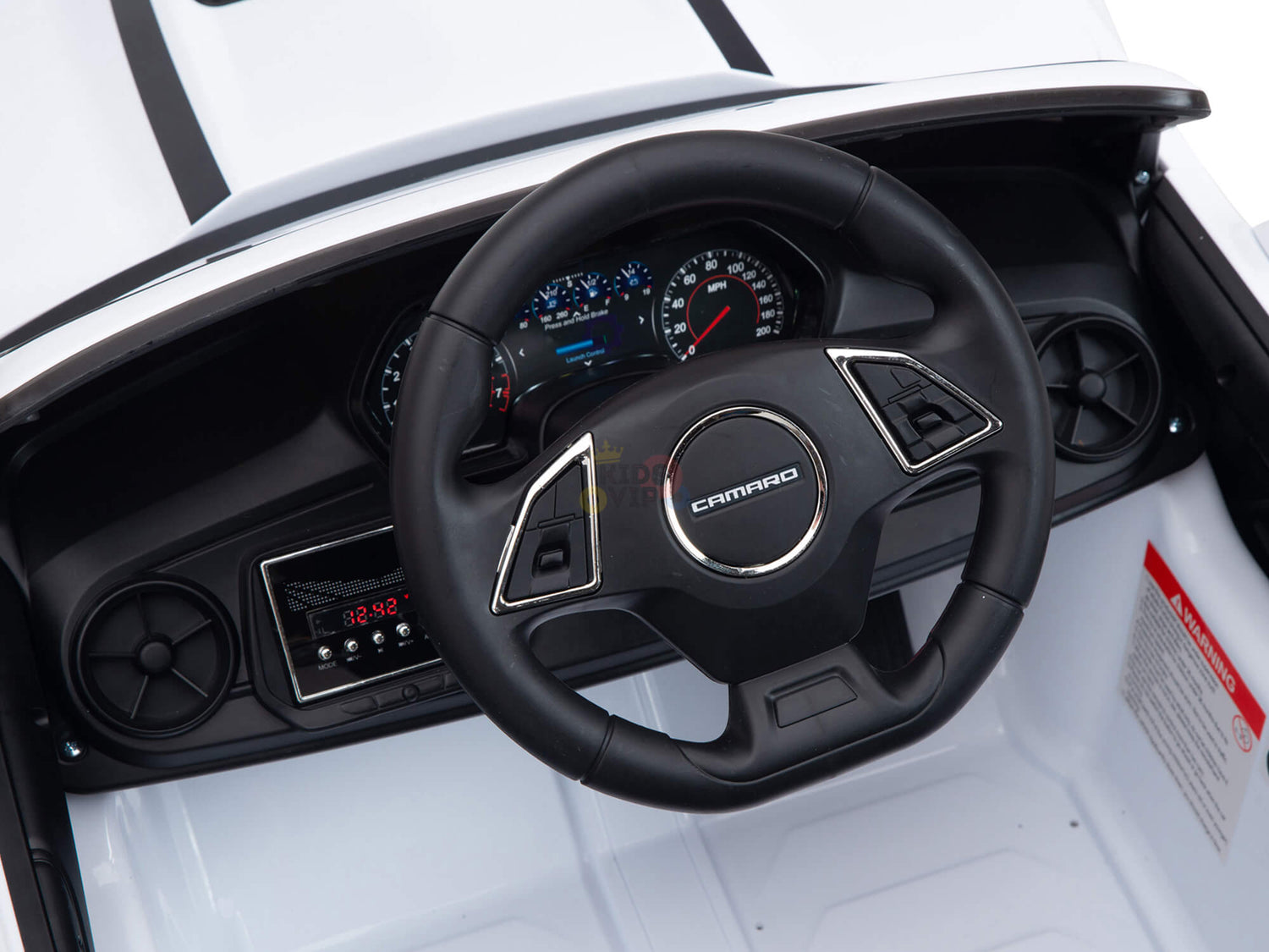 Close-up of the White 12V Official Sport Chevrolet Camaro Kids and Toddlers Ride on Car dashboard, showcasing a large black steering wheel with silver accents. A digital display behind the wheel shows speedometer and gauges, while the sleek black and white dash includes a digital radio screen for any remote control ride adventure.