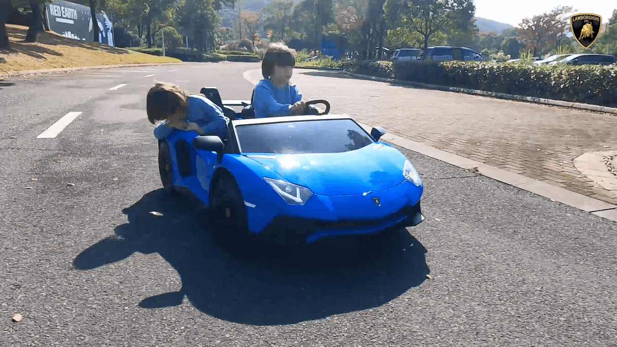 Two young children enjoy an exhilarating ride in their Blue Giant Official Limited Edition Lamborghini SV 24V/180W, zipping down a sunlit road at speeds of up to 10MPH. One child takes the eco-leather drivers seat while the other leans out, surrounded by trees and parked cars in a scenic backdrop.