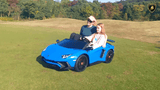 A woman in a hat and sunglasses smiles as a child steers the Blue Giant Official Limited Edition Lamborghini SV, a vibrant blue ride-on with eco-leather seats and a 180W motor. They cruise at up to 10MPH past trees and hills on a grassy field.