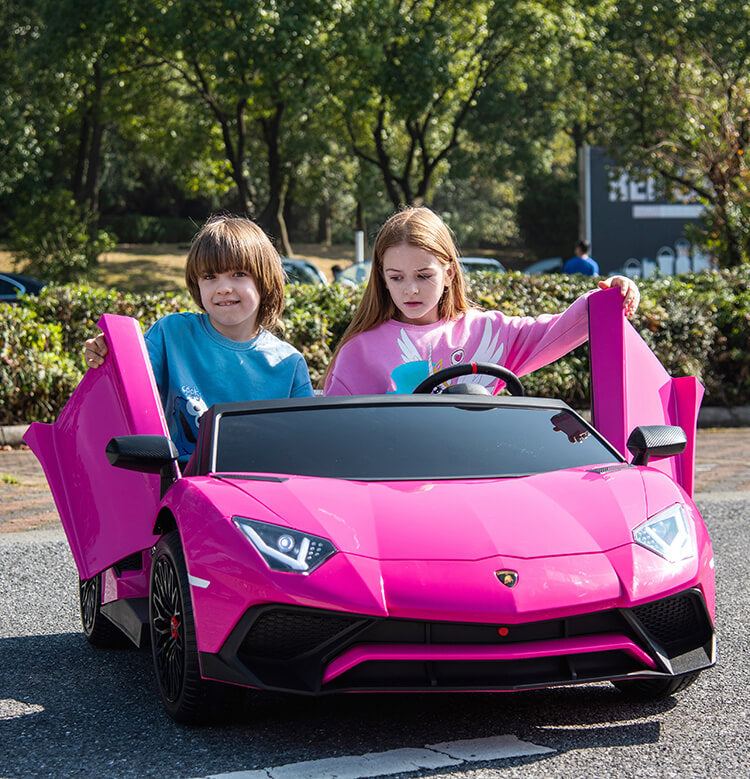 Two kids sit smiling in a Pink 24V/180W Lamborghini SV Limited XXL Edition ride-on, doors open. Parked on a paved road with trees and parking in the background, they enjoy this Kids VIP adventure.