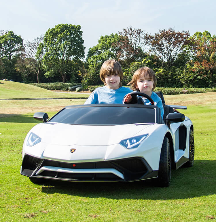 Two kids sit in a White 24V/180W Lamborghini SV Limited XXL Edition on a grassy field. The smiling child on the left, and the one holding the steering wheel on the right enjoy clear skies and trees in view, with speeds up to 10MPH.