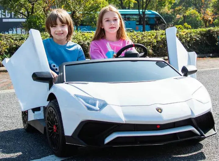 Two children enjoy a sunny day in their White 24V/180W Lamborghini SV Limited XXL Edition, with open scissor doors. The child on the left wears a blue shirt, and the one on the right sports pink, both seated comfortably on eco-leather seats, savoring their adventure at up to 10MPH outdoors.