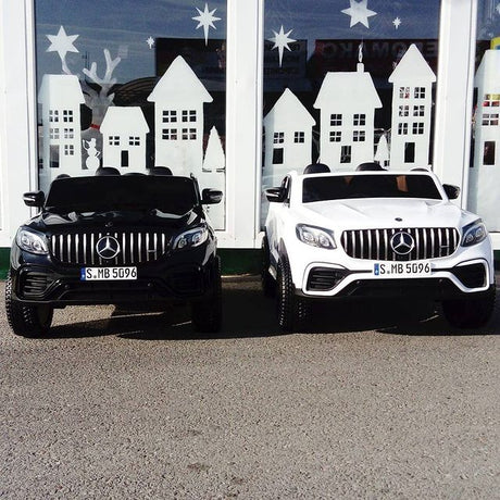 Two sleek 2-seater Mercedes GLC ride-on cars, one black and one white, parked on a pavement in front of a large window with white house and star cut-out designs, resemble toys for enthusiasts.