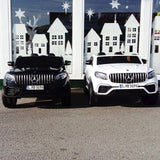 Two toy Mercedes-Benz cars, one black and one white, are parked side by side on a paved surface in front of a shop window with white house and star silhouettes, showcasing the remote-controlled performance of the Enhance Your Ride with a Remote Control for 2 Seater Mercedes GLC.